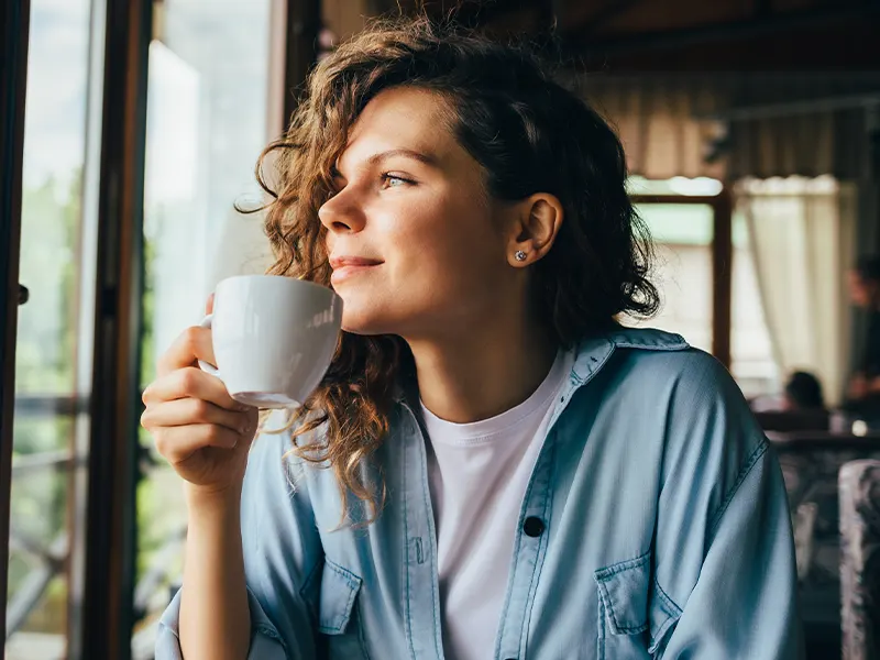 mujer tomando café en taza blanca mientras sonríe y mira por la ventana